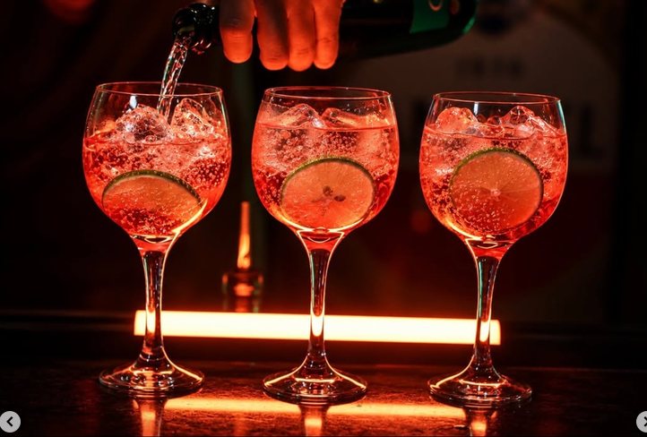 Três taças de drink avermelhado com gelo e limão, sendo servidas no balcão iluminado